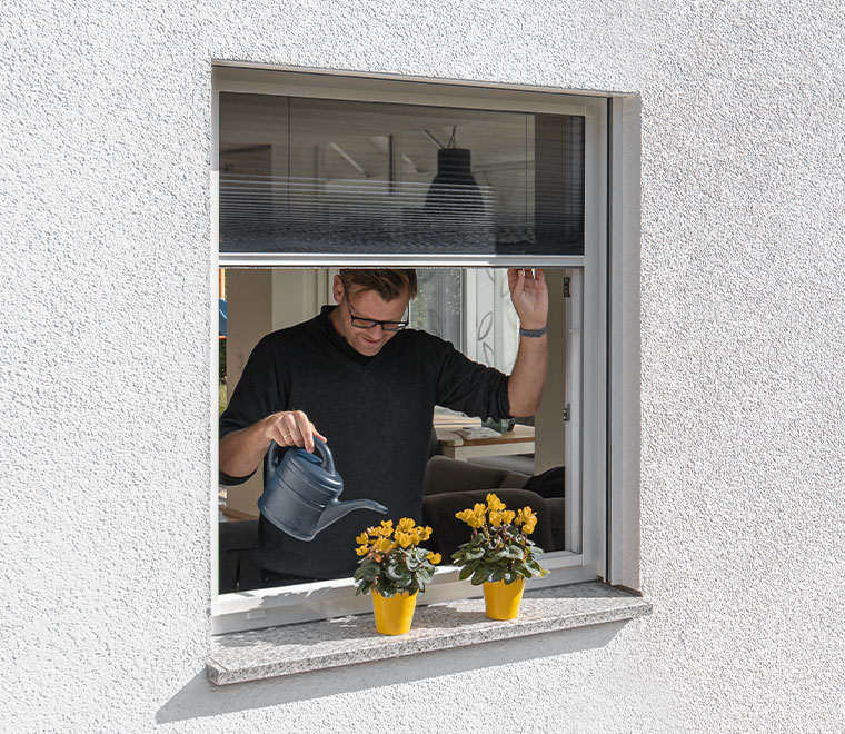 fliegengitter-rollo Ein Mann bewässert Pflanzen im Fenster, das mit einem Fliegengitter-Rollo geschützt ist.
