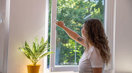 Frau in weißem T-Shirt öffnet ein Fenster, im Vordergrund eine Zimmerpflanze in gelbem Topf
